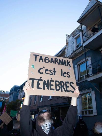Un manifestant·e avec un keffieh, une capuche, et le visage masqué sur photoshop brandit une pancarte : Tabarnak c'est les ténèbres
